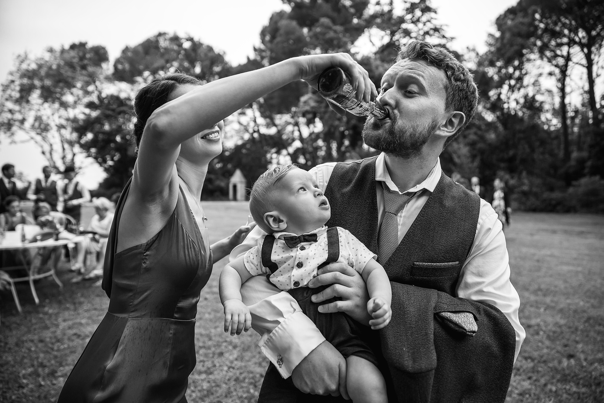 photojournalistic wedding photography of a man holding a baby while his wife holds up a beer bottle and gets him to drink beer, the baby watches the man drink. 