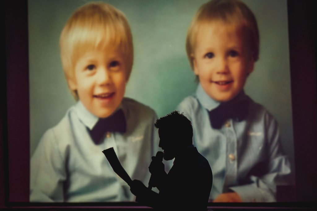 The brother of the groom gives a speech about him and the groom, their child hood pictures are on a screen and he is in silhouette. It is a documentary wedding photograph.